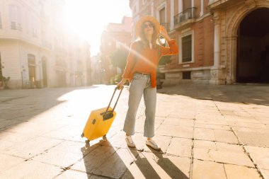 Şapka takan ve sarı bir bavul taşıyan bir turist gün batımında şehir sokaklarında geziniyor. Genç bir kadın bavulla seyahat etmekten hoşlanır. Konseptler: turizm, tatil, hafta sonu.