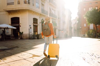 Şapka takan ve sarı bir bavul taşıyan bir turist gün batımında şehir sokaklarında geziniyor. Genç bir kadın bavulla seyahat etmekten hoşlanır. Konseptler: turizm, tatil, hafta sonu.
