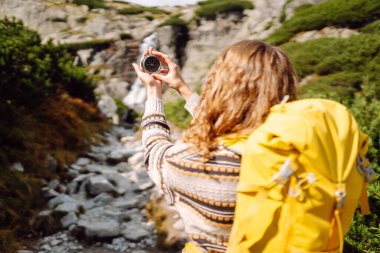 Sırt çantası ve pusula taşıyan kadın yürüyüşçünün arka plandaki kayalık patikada görüntüsü. Yürüyüşçü varış noktasını arar ve güneşli bir günde dağ manzarasının tadını çıkarır. Doğa kavramları, navigasyon.