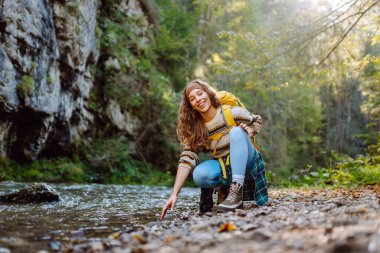 Sarı sırt çantalı bir kadın yürüyüşçü ormanla çevrili dağ nehrinin yanındaki suya dokunur. Güneşli bir günde ormanda yürüyüş parkurunda güzel bir kadın. Yürüyüş, macera, doğa kavramları.