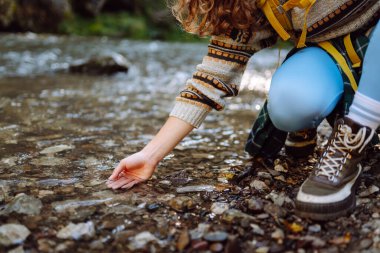 Sarı sırt çantalı bir kadın yürüyüşçü ormanla çevrili dağ nehrinin yanındaki suya dokunur. Güneşli bir günde ormanda yürüyüş parkurunda güzel bir kadın. Yürüyüş, macera, doğa kavramları.