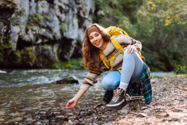 Sarı sırt çantalı bir kadın yürüyüşçü ormanla çevrili dağ nehrinin yanındaki suya dokunur. Güneşli bir günde ormanda yürüyüş parkurunda güzel bir kadın. Yürüyüş, macera, doğa kavramları.
