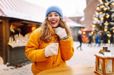 Parlak ceketli neşeli bir kadın atmosferik kış pazarında sıcak bir içecek tutuyor. Genç bir kadın turist, Noel öncesinde bir şehir pazarının keyfini çıkarıyor. Konsept şenlikli, rahat, sıcak içecekler..