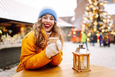 Parlak ceketli neşeli bir kadın atmosferik kış pazarında sıcak bir içecek tutuyor. Genç bir kadın turist, Noel öncesinde bir şehir pazarının keyfini çıkarıyor. Konsept şenlikli, rahat, sıcak içecekler..