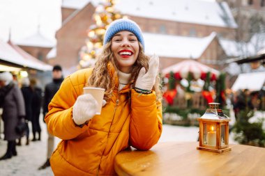 Parlak ceketli neşeli bir kadın atmosferik kış pazarında sıcak bir içecek tutuyor. Genç bir kadın turist, Noel öncesinde bir şehir pazarının keyfini çıkarıyor. Konsept şenlikli, rahat, sıcak içecekler..
