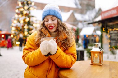 Parlak ceketli neşeli bir kadın atmosferik kış pazarında sıcak bir içecek tutuyor. Genç bir kadın turist, Noel öncesinde bir şehir pazarının keyfini çıkarıyor. Konsept şenlikli, rahat, sıcak içecekler..