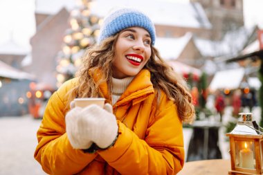 Parlak ceketli neşeli bir kadın atmosferik kış pazarında sıcak bir içecek tutuyor. Genç bir kadın turist, Noel öncesinde bir şehir pazarının keyfini çıkarıyor. Konsept şenlikli, rahat, sıcak içecekler..