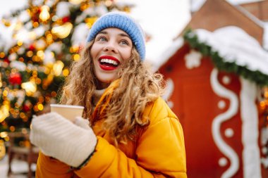 Parlak ceketli neşeli bir kadın atmosferik kış pazarında sıcak bir içecek tutuyor. Genç bir kadın turist, Noel öncesinde bir şehir pazarının keyfini çıkarıyor. Konsept şenlikli, rahat, sıcak içecekler..