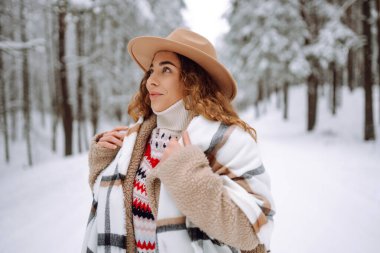 Kar ormanlarında gezen şapkalı gülümseyen bir kadının portresi. Sıcak ve şık giyinmiş güzel bir kadın ormanda güneşli bir kış gününün tadını çıkarıyor. Tarz, güzellik ve doğa kavramı.