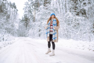 Neşeli bir kadın, kalın giysiler içinde, karlı bir yolda duruyor. Şapkalı bir yürüyüşçü ormanda karlı bir günün tadını çıkarır. Doğa, yürüyüş ve yaşam tarzı kavramları.