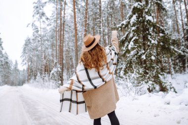 Neşeli bir kadın, kalın giysiler içinde, karlı bir yolda duruyor. Şapkalı bir yürüyüşçü ormanda karlı bir günün tadını çıkarır. Doğa, yürüyüş ve yaşam tarzı kavramları.