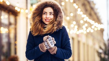 Mavi ceketli, örgü şapkalı ve eldivenli güzel bir kız şehrin caddesinde poz veriyor. Çelenk ışıkları.