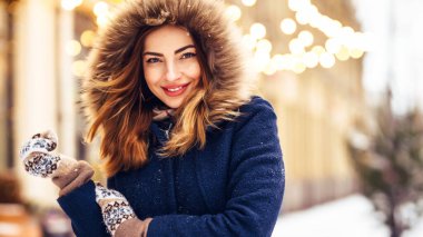 Mavi ceketli, örgü şapkalı ve eldivenli güzel bir kız şehrin caddesinde poz veriyor. Çelenk ışıkları.