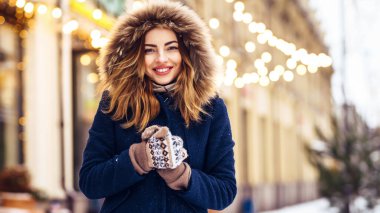 Mavi ceketli, örgü şapkalı ve eldivenli güzel bir kız şehrin caddesinde poz veriyor. Çelenk ışıkları.