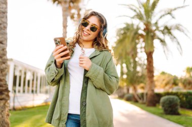 Kıvırcık saçlı, kablosuz kulaklık takan ve telefonunu kullanan neşeli bir kadın parkta güneşli bir günün tadını çıkarıyor. Güzel bir kadın müzik dinler ve telefona bakar. Yürüyüş kavramı ve hafta sonları..