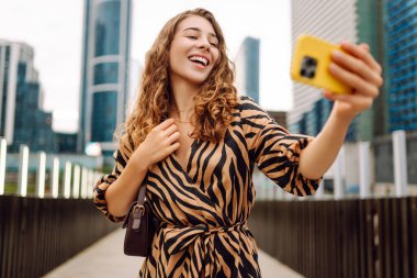 Selfie zamanı! Genç bir kadın şehrin hareketli şehirlerinde telefonla eğleniyor. Güzel turistler gezintiden hoşlanır, güneşli bir günde bloglar metropolün arka planına karşı. Bloglama, kentsel, turizm kavramları.