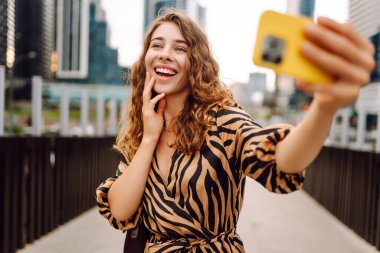 Selfie zamanı! Genç bir kadın şehrin hareketli şehirlerinde telefonla eğleniyor. Güzel turistler gezintiden hoşlanır, güneşli bir günde bloglar metropolün arka planına karşı. Bloglama, kentsel, turizm kavramları.