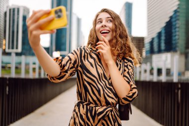 Selfie zamanı! Genç bir kadın şehrin hareketli şehirlerinde telefonla eğleniyor. Güzel turistler gezintiden hoşlanır, güneşli bir günde bloglar metropolün arka planına karşı. Bloglama, kentsel, turizm kavramları.