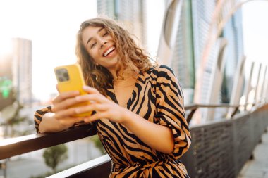 Neşeli bir kadın, elinde telefonla kalabalık bir metropolün arka planına poz veriyor. Güzel bir kadın blogları ve güneşli şehir sokaklarında geziniyor. Blog konsepti, yürümek. Yaşam biçimi.