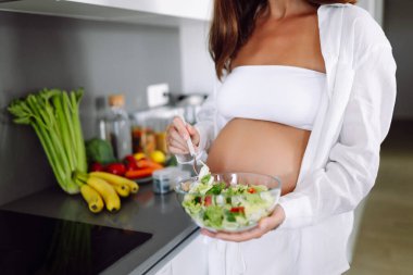 Bebek bekleyen güzel bir kadın şık mutfağında duruyor ve salatayı kaseye koyuyor. Hamile bir kadın öğle yemeğinden önce sıcak bir odada vakit geçirmekten hoşlanır. Sağlıklı beslenme, rahatlık ve beklenti kavramı.