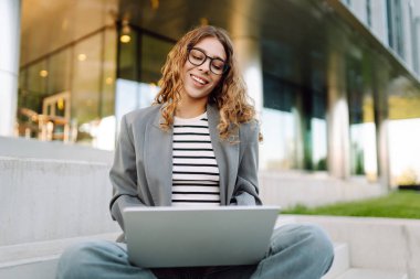 Gözlüklü genç bir kadın şık bir binanın önündeki basamaklarda dizüstü bilgisayarla oturuyor. Güneşli bir günde kadın bir serbest çalışan dışarıda çalışır. Uzak çalışma, teknoloji ve moda kavramları.