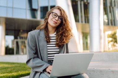 Gözlüklü genç bir kadın şık bir binanın önündeki basamaklarda dizüstü bilgisayarla oturuyor. Güneşli bir günde kadın bir serbest çalışan dışarıda çalışır. Uzak çalışma, teknoloji ve moda kavramları.