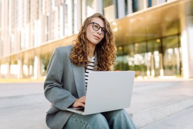 Gözlüklü genç bir kadın şık bir binanın önündeki basamaklarda dizüstü bilgisayarla oturuyor. Güneşli bir günde kadın bir serbest çalışan dışarıda çalışır. Uzak çalışma, teknoloji ve moda kavramları.