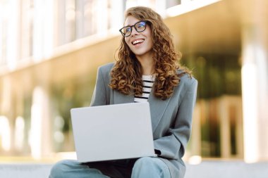 Gözlüklü genç bir kadın şık bir binanın önündeki basamaklarda dizüstü bilgisayarla oturuyor. Güneşli bir günde kadın bir serbest çalışan dışarıda çalışır. Uzak çalışma, teknoloji ve moda kavramları.