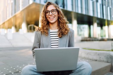 Gözlüklü genç bir kadın şık bir binanın önündeki basamaklarda dizüstü bilgisayarla oturuyor. Güneşli bir günde kadın bir serbest çalışan dışarıda çalışır. Uzak çalışma, teknoloji ve moda kavramları.