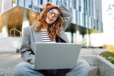 Gözlüklü genç bir kadın şık bir binanın önündeki basamaklarda dizüstü bilgisayarla oturuyor. Güneşli bir günde kadın bir serbest çalışan dışarıda çalışır. Uzak çalışma, teknoloji ve moda kavramları.