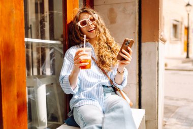 Şık giyinmiş gülümseyen bir kadın elinde soğuk bir içecekle bir kahve dükkanının yanındaki bankta oturur. Genç bir kadın, elinde telefon ve içkiyle güneşli bir günün tadını çıkarır. Konsept: İçkiler, rahatlama.