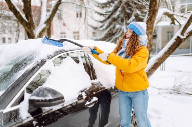 Parlak ceketli bir kadın sabah karlı bir bölgede arabanın ön camından kar temizliyor. Kadın sürücü güneşli kış gününde arabanın camından karı temizler. Ulaşım, temizlik ve mevsimlik kavramları.