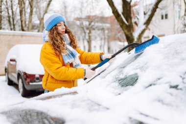 Parlak ceketli bir kadın sabah karlı bir bölgede arabanın ön camından kar temizliyor. Kadın sürücü güneşli kış gününde arabanın camından karı temizler. Ulaşım, temizlik ve mevsimlik kavramları.