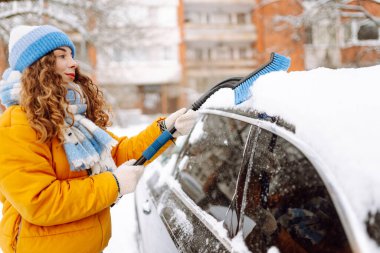 Parlak ceketli bir kadın sabah karlı bir bölgede arabanın ön camından kar temizliyor. Kadın sürücü güneşli kış gününde arabanın camından karı temizler. Ulaşım, temizlik ve mevsimlik kavramları.