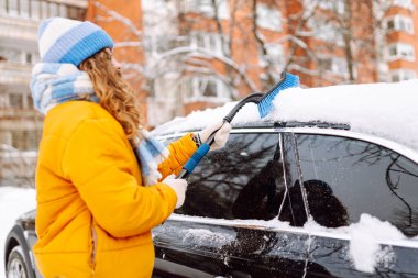 Parlak ceketli bir kadın sabah karlı bir bölgede arabanın ön camından kar temizliyor. Kadın sürücü güneşli kış gününde arabanın camından karı temizler. Ulaşım, temizlik ve mevsimlik kavramları.