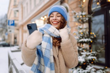 Karlı şehir caddesinde telefonunu kullanan kıvırcık saçlı genç bir kadının portresi. Parlak atkı ve şapkalı neşeli kadın güneşli kış gününde mesaj atıyor ya da blog yazıyor. Kavram: Kentsel, blog yazarlığı. Yaşam biçimi.