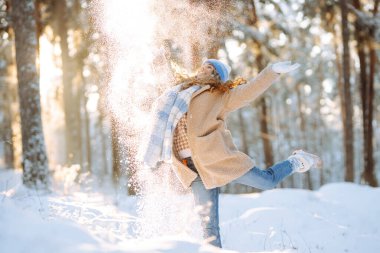 Sıcak kıyafetler giymiş neşeli bir kadın güneşli bir ormanda karla eğleniyor. Güzel bir kadın doğayı seviyor, kış günü açık havada havaya kar atıyor. Mevsimlik, kar, keyif kavramları.