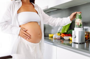Hamile kadın modern mutfakta duruyor, cama yeşil smoothie döküyor. Müstakbel anne sıcak uzayda taze sebze ve meyvelerle tezgahın yanında duruyor. Sağlık ve içecek kavramı..