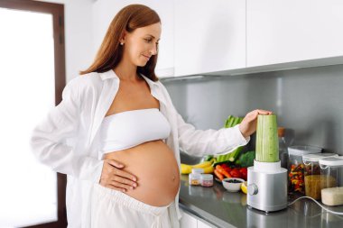 Hamile kadın modern mutfakta duruyor, cama yeşil smoothie döküyor. Müstakbel anne sıcak uzayda taze sebze ve meyvelerle tezgahın yanında duruyor. Sağlık ve içecek kavramı..