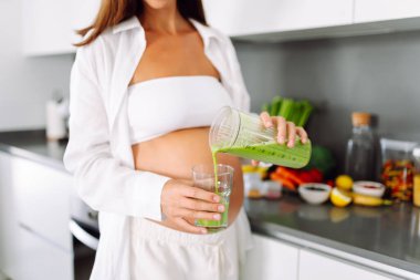 Hamile kadın modern mutfakta duruyor, cama yeşil smoothie döküyor. Müstakbel anne sıcak uzayda taze sebze ve meyvelerle tezgahın yanında duruyor. Sağlık ve içecek kavramı..