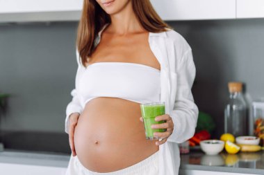 Hamile kadın modern mutfakta duruyor, cama yeşil smoothie döküyor. Müstakbel anne sıcak uzayda taze sebze ve meyvelerle tezgahın yanında duruyor. Sağlık ve içecek kavramı..
