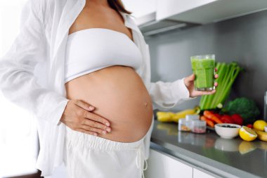 Hamile kadın modern mutfakta duruyor, cama yeşil smoothie döküyor. Müstakbel anne sıcak uzayda taze sebze ve meyvelerle tezgahın yanında duruyor. Sağlık ve içecek kavramı..