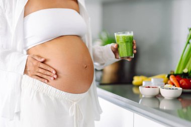 Hamile kadın modern mutfakta duruyor, cama yeşil smoothie döküyor. Müstakbel anne sıcak uzayda taze sebze ve meyvelerle tezgahın yanında duruyor. Sağlık ve içecek kavramı..