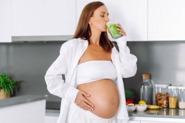 Hamile kadın modern mutfakta duruyor, cama yeşil smoothie döküyor. Müstakbel anne sıcak uzayda taze sebze ve meyvelerle tezgahın yanında duruyor. Sağlık ve içecek kavramı..