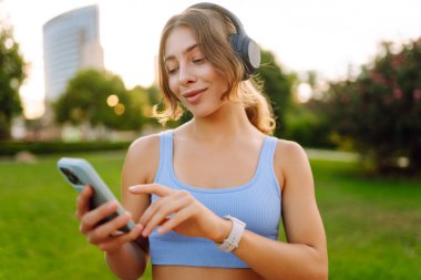 Güneşli bir parkta kablosuz kulaklık takan ve elinde telefon tutan genç bir kadın. Spor giyim sektöründe bir kadın açık havada müzik dinlemekten hoşlanır. Zevk kavramı, teknoloji.