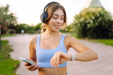 Güneşli bir parkta kablosuz kulaklık takan ve elinde telefon tutan genç bir kadın. Spor giyim sektöründe bir kadın açık havada müzik dinlemekten hoşlanır. Zevk kavramı, teknoloji.