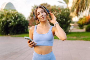 Güneşli bir parkta kablosuz kulaklık takan ve elinde telefon tutan genç bir kadın. Spor giyim sektöründe bir kadın açık havada müzik dinlemekten hoşlanır. Zevk kavramı, teknoloji.