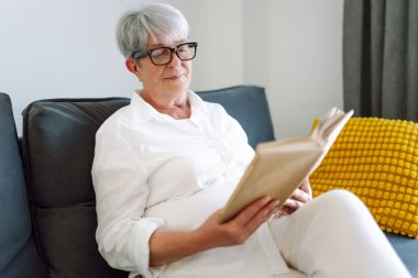 Yaşlı bir kadın rahat bir oturma odasında kanepede oturup kitap okuyor. Gri saçlı kadın kitap okumayı ve odada dinlenmeyi sever. Mutlu yaşlanma, rahatlama ve rahatlama kavramı..