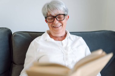 Yaşlı bir kadın rahat bir oturma odasında kanepede oturup kitap okuyor. Gri saçlı kadın kitap okumayı ve odada dinlenmeyi sever. Mutlu yaşlanma, rahatlama ve rahatlama kavramı..