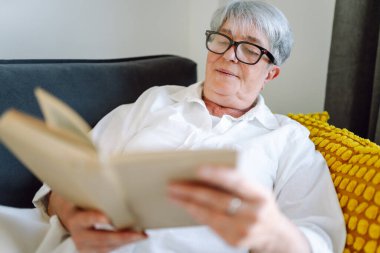 Yaşlı bir kadın rahat bir oturma odasında kanepede oturup kitap okuyor. Gri saçlı kadın kitap okumayı ve odada dinlenmeyi sever. Mutlu yaşlanma, rahatlama ve rahatlama kavramı..